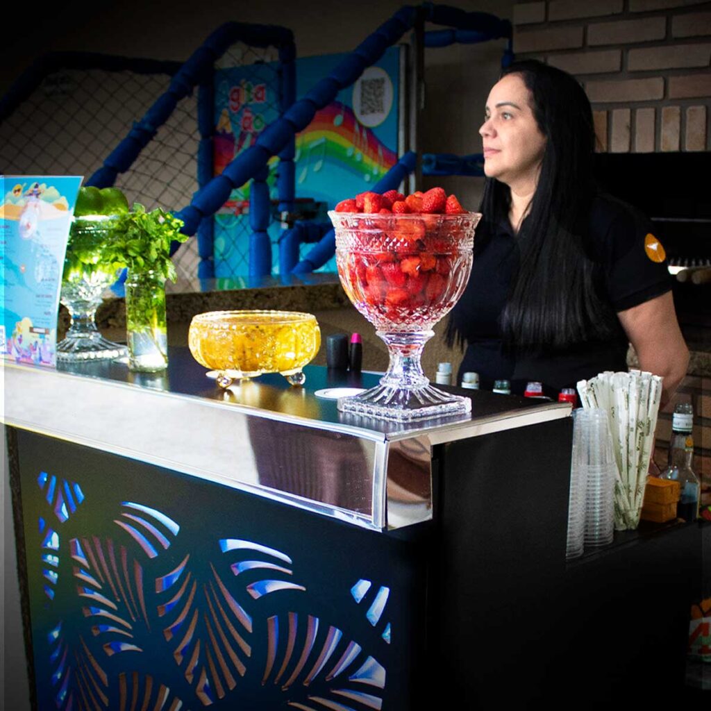 Bartender de prontidão para iniciar os serviços de bar em uma festa de aniversário. Ela está atrás do bar. Em cima do balcão tem 3 fruteiras e hortelã para montar caipirinhas e Mojito. As frutas são Morango, Maracujá e Limão taiti, será servido com vodka, saque ou cachaça