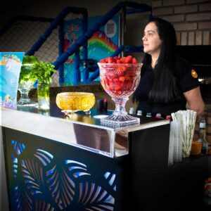 Bartender de prontidão para iniciar os serviços de bar em uma festa de aniversário. Ela está atrás do bar. Em cima do balcão tem 3 fruteiras e hortelã para montar caipirinhas e Mojito. As frutas são Morango, Maracujá e Limão taiti, será servido com vodka, saque ou cachaça