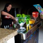 Bartender preparando um drink delicioso chamado beijo na boca. Ela está segurando uma garrafa de xarope de romã e finalizando o drink para entregar ao convidado que está a sua frente segurando o cardápio. Dentre o convidado e a Bartender há uma estrutura de bar, e em cima do balcão tem 3 tipos de frutas para o festival de caipirinhas, a primeira fruteira é limão taiti, segunda é hortelã para preparar mojito, em sequência tem uma compoteira de cristal cheia de maracujá e por fim tem outra fruteira cheia de morango para fazer caipiroskas de morango ou saquerinhas de morango.