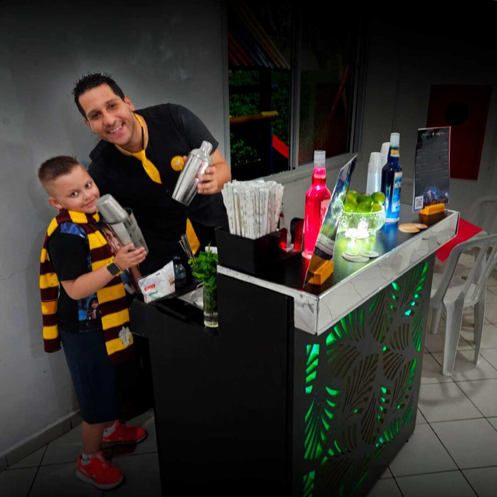 Barman sorridente com criança em festa em São Paulo, ambos segurando coqueteleiras em frente a um bar iluminado.