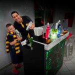 Barman sorridente com criança em festa em São Paulo, ambos segurando coqueteleiras em frente a um bar iluminado.