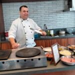 Chef de buffet de crepe para festas preparando crepes na chapa com diversos recheios salgados organizados em recipientes de inox.