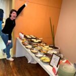 Mulher sorridente ao lado de uma mesa completa de buffet de crepe para festas, mostrando a variedade de recheios, pratos e o serviço de crepes sendo preparados.