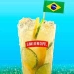 Copo de caipiroska com vodka Smirnoff bem gelada, repleto de fatias de limão siciliano, limão taiti e gelo picado, decorado com uma pequena bandeira do Brasil no topo, contra um fundo azul brilhante.