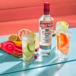 Garrafa de Vodka Smirnoff 998ml no cento. Ao lado direito 2 copos alto com caipirinhas de grapefruit e a outra de limão siciliano. Ao lado esquerdo outro copo com caipiroska de limão taiti. Ao fundo tem uma tábua com as frutas que o Bartender cortou para fazer as 3 caipiroskas.