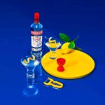 Garrafa de Vodka Smirnoff 998ml no fundo lado esquerdo, 2 copos cônicos com vodka e guarnição, um está em cima de uma tábua circulas amarela e o outro está fora ao lado de um descascador amarelo. Em cima da tábua tem um dosados e um limão siciliano
