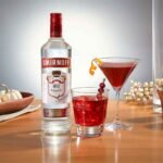 Garrafa de vodka Smirnoff No 21 original ao lado de dois coquetéis vermelhos sobre uma mesa de madeira iluminada, sendo um drink em copo baixo com gelo e mirtilos, e outro em taça martini decorado com casca de laranja.