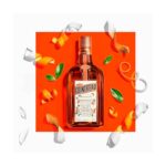 Garrafa de licor Cointreau em fundo laranja com cascas de laranja e folhas verdes ao redor.