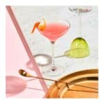 Coquetel Cosmopolitan rosa servido em elegante taça coupe com garnish de laranja, ao lado de acessórios de bar.