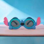 Óculos infantis do Stitch em primeiro plano sobre uma superfície rosa e um fundo azul claro.
