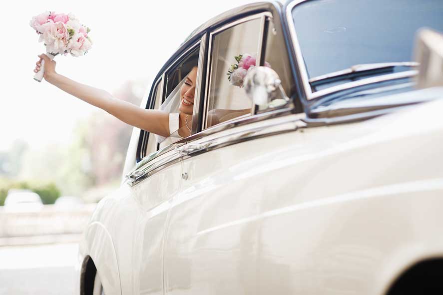 Noiva feliz com vestido branco acenando com um buquê de flores cor-de-rosa pela janela de um carro clássico branco. A noiva está sorrindo.