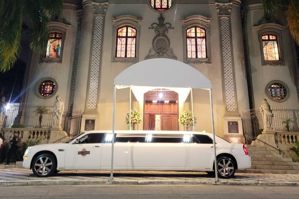 Limousine branca longa estacionada em frente a uma igreja iluminada à noite. Palavra-chave: limousine para casamento em São Paulo.