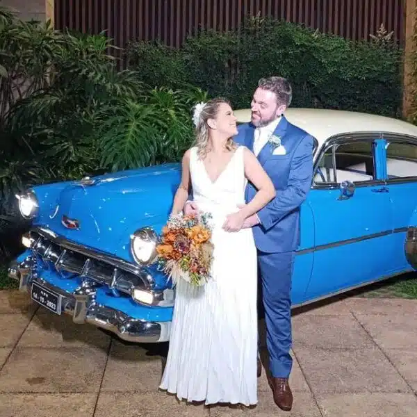 Casal de noivos em frente a um carro antigo azul e branco, possivelmente um Chevrolet de época. A noiva está de vestido branco com um buquê e o noivo de terno azul, abraçando-a.