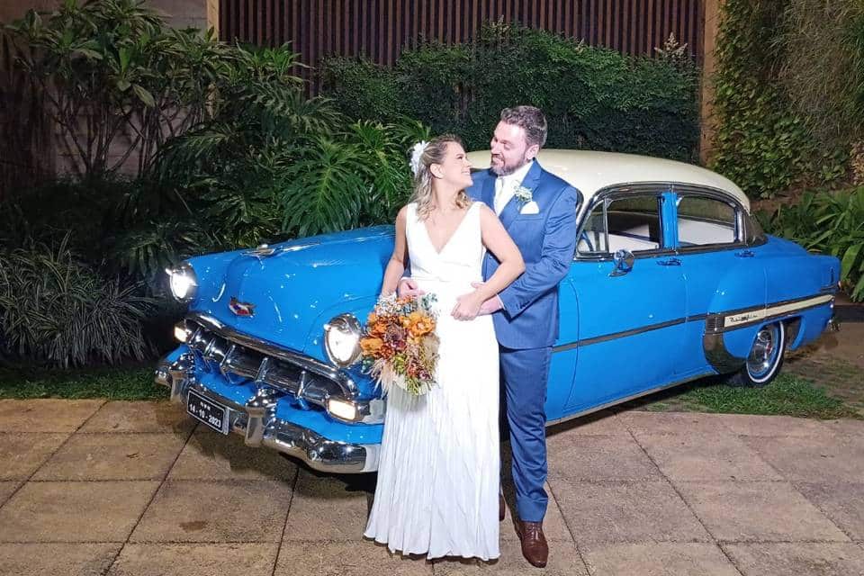 Casal de noivos em frente a um carro antigo azul e branco, possivelmente um Chevrolet de época. A noiva está de vestido branco com um buquê e o noivo de terno azul, abraçando-a.