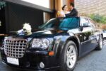 alt: Casal de noivos em um Chrysler 300 preto brindando com taças de champanhe. A noiva está de vestido branco e o noivo de terno escuro. Há um buquê branco no capô do carro. Palavra-chave: transporte executivo para casamento em São Paulo.