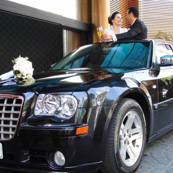 alt: Casal de noivos em um Chrysler 300 preto brindando com taças de champanhe. A noiva está de vestido branco e o noivo de terno escuro. Há um buquê branco no capô do carro. Palavra-chave: transporte executivo para casamento em São Paulo.