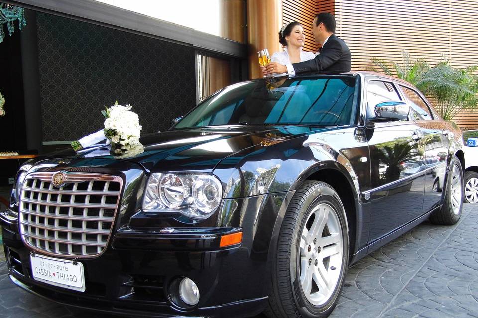 alt: Casal de noivos em um Chrysler 300 preto brindando com taças de champanhe. A noiva está de vestido branco e o noivo de terno escuro. Há um buquê branco no capô do carro. Palavra-chave: transporte executivo para casamento em São Paulo.