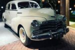 Carro retrô creme ou bege claro, com para-choques cromados e pneus de banda branca, estacionado em ambiente externo com iluminação aconchegante. Palavra-chave: carros retrô para casamento em São Paulo.