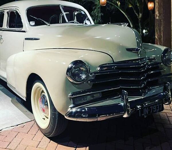Carro retrô creme ou bege claro, com para-choques cromados e pneus de banda branca, estacionado em ambiente externo com iluminação aconchegante. Palavra-chave: carros retrô para casamento em São Paulo.