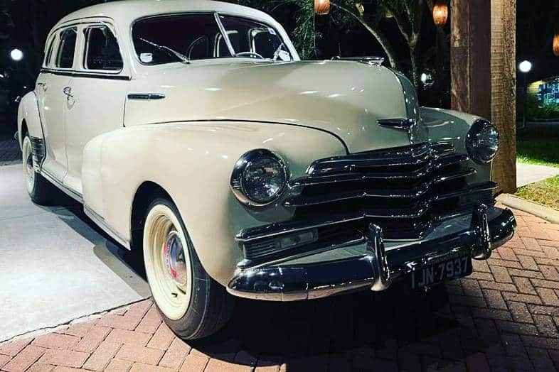 Carro retrô creme ou bege claro, com para-choques cromados e pneus de banda branca, estacionado em ambiente externo com iluminação aconchegante. Palavra-chave: carros retrô para casamento em São Paulo.