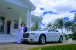 Noivos em frente a uma casa branca com um carro de luxo branco, um Chrysler 300, estacionado. A noiva está de vestido branco e o noivo de terno azul. Palavra-chave: carro de luxo casamento São Paulo.