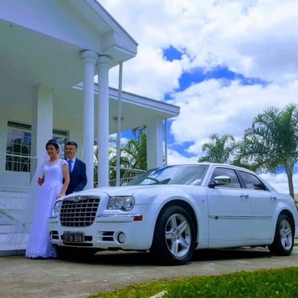Noivos em frente a uma casa branca com um carro de luxo branco, um Chrysler 300, estacionado. A noiva está de vestido branco e o noivo de terno azul. Palavra-chave: carro de luxo casamento São Paulo.
