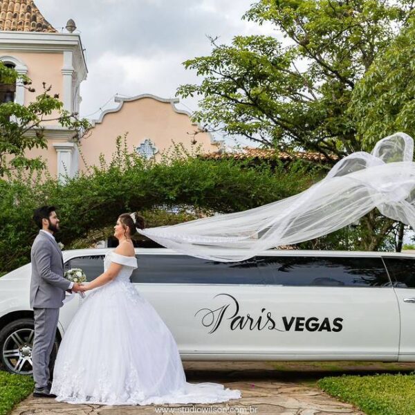Casal de noivos em frente a uma limousine branca de luxo com o logo "Paris Vegas". O véu da noiva flutua ao vento.