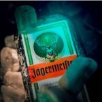Close-up em ângulo inclinado de uma garrafa de licor Jägermeister extremamente congelada, cercada por cubos de gelo sob uma iluminação azul e esverdeada estilo neon que destaca o rótulo do cervo.