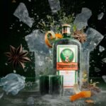 Garrafa fosca e congelada de licor Jägermeister cercada por cubos de gelo a explodir, raspas de laranja, anis-estrelado e botânicos, com dois copos de shot personalizados à frente.
